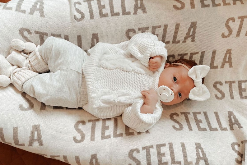 May include: A baby wearing a white knitted sweater and white pants lies on a cream-colored blanket with the name "Stella" repeated in brown lettering.