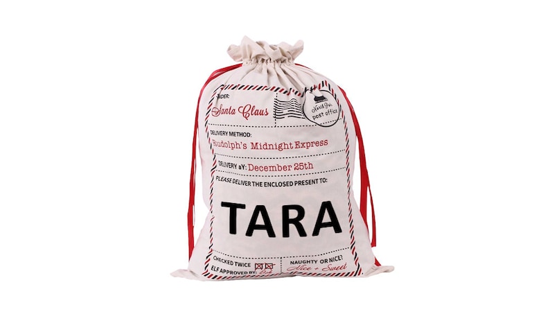 May include: White canvas Santa sack with red ribbon ties. The sack has a printed label that says "Santa Claus" and "Rudolph's Midnight Express". The label also says "Delivery by: December 25th" and "Please deliver the enclosed present to: TARA".