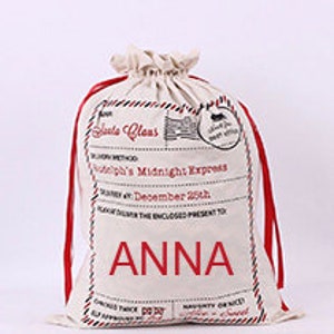 May include: A white canvas Santa sack with red ribbon ties. The sack has a vintage-style label that reads "Santa Claus" and "Rudolph's Midnight Express". The name "ANNA" is printed in large letters on the front of the sack.