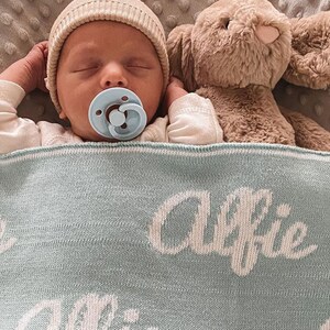 May include: A baby wearing a beige knitted hat and a blue pacifier sleeps on a light blue knitted blanket with the name "Alfie" embroidered in white. A brown plush bunny is beside the baby.