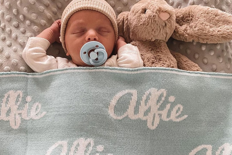 May include: A baby wearing a beige knitted hat and a blue pacifier sleeps on a light blue knitted blanket with the name "Alfie" embroidered in white. A brown plush bunny is beside the baby.