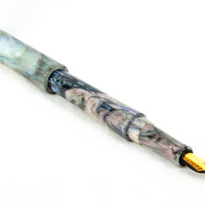 K&ouml;nnte beinhalten: Ein marmorierter F&uuml;llfederhalter mit goldener Feder. Der Stift hat ein wirbelndes Muster aus Blau, Grau und Wei&szlig;. Kappe und Korpus haben eine gl&auml;nzende Oberfl&auml;che. Die Feder ist goldfarben.