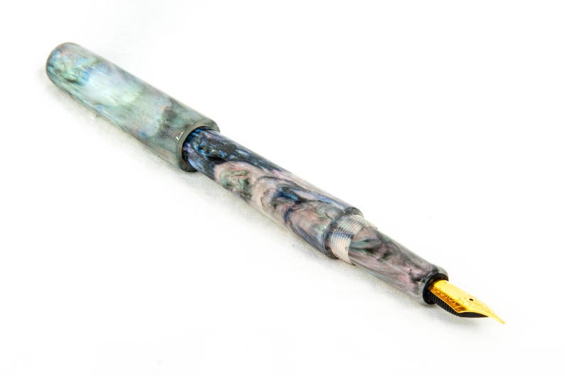 K&ouml;nnte beinhalten: Ein marmorierter F&uuml;llfederhalter mit goldener Feder. Der Stift hat ein wirbelndes Muster aus Blau, Grau und Wei&szlig;. Kappe und Korpus haben eine gl&auml;nzende Oberfl&auml;che. Die Feder ist goldfarben.