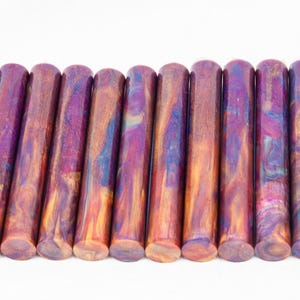 May include: A row of 15 colorful, iridescent acrylic pen blanks. The blanks are a mix of purple, blue, gold, and orange colors. The blanks are all the same size and shape, and they are arranged in a row on a white background.