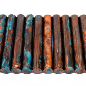 May include: A collection of twelve cylindrical wooden rods. Each rod features a swirling pattern of brown, teal, and orange hues. The rods are arranged horizontally against a white background, showcasing their unique color variations and textures.