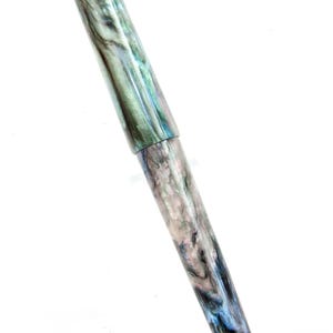 K&ouml;nnte beinhalten: Ein F&uuml;llfederhalter mit einem marmorierten Design in Gr&uuml;n-, Blau- und Graut&ouml;nen. Der Stift hat eine Kappe und einen schlanken, l&auml;nglichen K&ouml;rper. Der Stift wird vor einem wei&szlig;en Hintergrund pr&auml;sentiert.