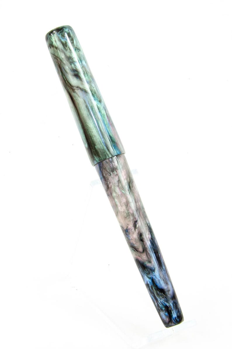 K&ouml;nnte beinhalten: Ein F&uuml;llfederhalter mit einem marmorierten Design in Gr&uuml;n-, Blau- und Graut&ouml;nen. Der Stift hat eine Kappe und einen schlanken, l&auml;nglichen K&ouml;rper. Der Stift wird vor einem wei&szlig;en Hintergrund pr&auml;sentiert.