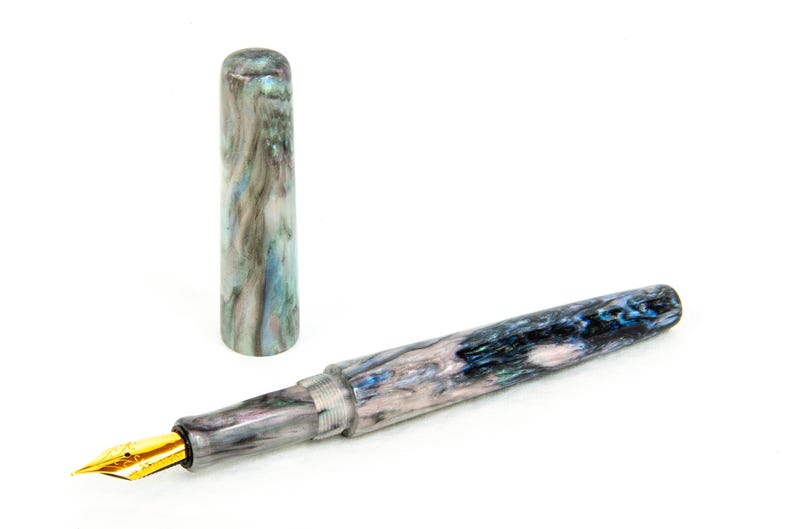 K&ouml;nnte beinhalten: Ein F&uuml;llfederhalter mit marmoriertem Design in Blau-, Grau- und Wei&szlig;t&ouml;nen, mit einer goldfarbenen Feder. Die Kappe ist abgenommen und neben den Stift gelegt. Der Stift befindet sich auf wei&szlig;em Hintergrund.