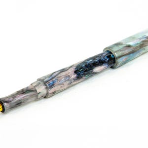 K&ouml;nnte beinhalten: Ein F&uuml;llfederhalter mit einem marmorierten, mehrfarbigen Schaft in Grau-, Blau- und Wei&szlig;t&ouml;nen. Der Stift hat eine goldfarbene Feder und einen silberfarbenen Metallring in der N&auml;he der Feder. Der Stift ist vor einem wei&szlig;en Hintergrund platziert.