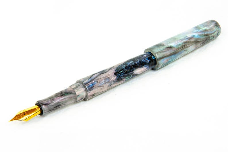 K&ouml;nnte beinhalten: Ein F&uuml;llfederhalter mit einem marmorierten, mehrfarbigen Schaft in Grau-, Blau- und Wei&szlig;t&ouml;nen. Der Stift hat eine goldfarbene Feder und einen silberfarbenen Metallring in der N&auml;he der Feder. Der Stift ist vor einem wei&szlig;en Hintergrund platziert.
