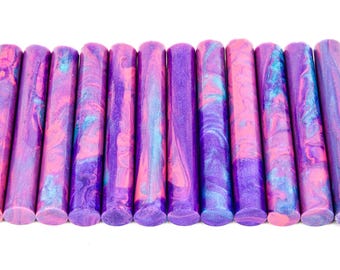 May include: A collection of eleven cylindrical wax sticks in shades of purple, pink, and blue. The wax sticks have a marbled appearance, with swirls of color throughout. The ends are flat and the sticks are arranged horizontally.