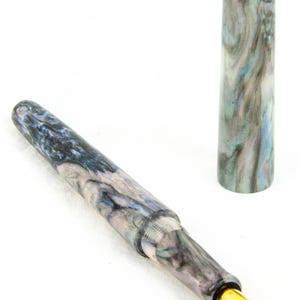 K&ouml;nnte beinhalten: Ein marmorierter F&uuml;llfederhalter mit goldener Feder und Kappe. Der Stiftk&ouml;rper weist ein wirbelndes Muster aus Blau, Grau und Wei&szlig; auf. Die Kappe ist abgenommen und steht aufrecht und zeigt das gleiche Marmormuster. Der Stift ist zum Schreiben bestimmt.