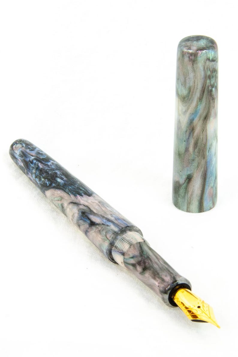 K&ouml;nnte beinhalten: Ein marmorierter F&uuml;llfederhalter mit goldener Feder und Kappe. Der Stiftk&ouml;rper weist ein wirbelndes Muster aus Blau, Grau und Wei&szlig; auf. Die Kappe ist abgenommen und steht aufrecht und zeigt das gleiche Marmormuster. Der Stift ist zum Schreiben bestimmt.