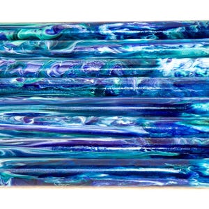 Puede incluir: Una pila de diez bloques cilíndricos para bolígrafos en tonos azules, turquesas y blancos. La resina presenta un patrón veteado y arremolinado, creando una apariencia única y artística. Los bloques están apilados horizontalmente.
