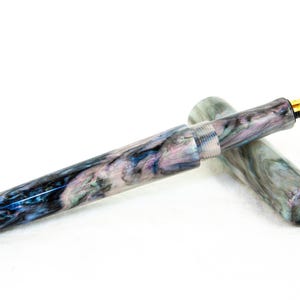 K&ouml;nnte beinhalten: Ein marmorierter F&uuml;llfederhalter mit goldener Feder. Der Stiftk&ouml;rper weist ein wirbelndes Muster aus Blau, Lila und Wei&szlig; auf. Die Kappe ist abgenommen und gibt die Feder frei. Der Stift ist ein Schreibger&auml;t.