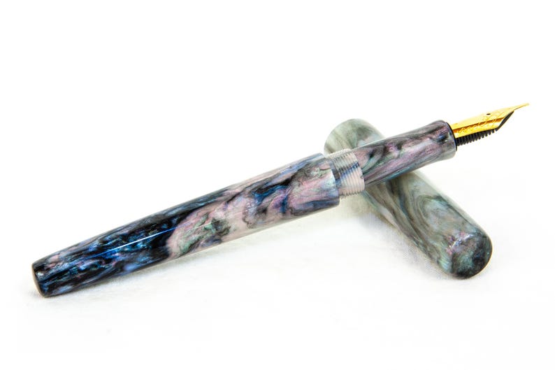 K&ouml;nnte beinhalten: Ein marmorierter F&uuml;llfederhalter mit goldener Feder. Der Stiftk&ouml;rper weist ein wirbelndes Muster aus Blau, Lila und Wei&szlig; auf. Die Kappe ist abgenommen und gibt die Feder frei. Der Stift ist ein Schreibger&auml;t.