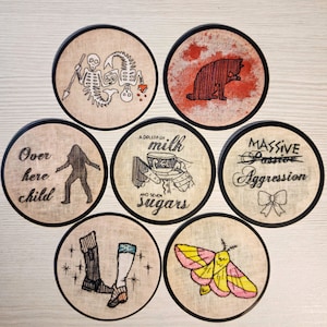 May include: Set of 7 round coasters with various designs. The coasters are made of a beige fabric with black trim. The designs include a skeleton, a cat, a moth, a pair of boots, a woman, a milk carton, and a bow with the text "MASSIVE Passive Aggression".