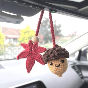 May include: Crocheted car charm featuring a red maple leaf and a smiling acorn with a brown cap. The charm is suspended by a red cord with a wooden bead, adding a touch of handmade charm to the car's interior.