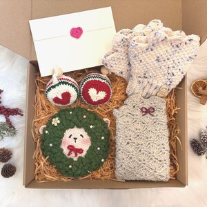 May include: A gift box filled with handmade crochet items. Includes a pair of speckled gloves, a matching arm warmer, two ornaments, a wreath, and a card with a pink heart. The box is lined with straw.