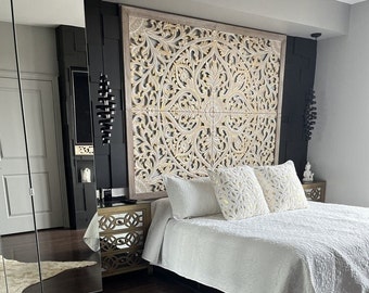 May include: A white bed with a white quilt and white pillows. The headboard is a large, intricate wooden carving with a light brown finish. The bed is in a bedroom with dark wood floors and a white rug.