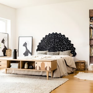 May include: A black wooden headboard featuring a large, intricate mandala design. The headboard is mounted on a white wall above a bed with a beige duvet and patterned pillows. The bed is in a bedroom with a wooden floor and a rug.
