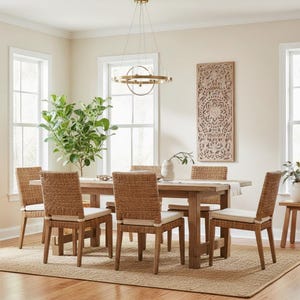 Può includere: Un tavolo da pranzo rettangolare in legno con sei sedie in rattan intrecciato dotate di cuscini color bianco sporco. Il set è disposto su un tappeto in juta testurizzato, completato da un lampadario circolare in metallo dorato e un pannello in legno.
