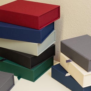 Peut inclure: Plusieurs boîtes cadeaux rectangulaires de différentes couleurs, dont rouge, bleu marine, gris, noir et vert. Les boîtes sont empilées et disposées sur une surface blanche, mettant en valeur leurs différentes teintes et textures. Certaines boîtes ont des rubans.