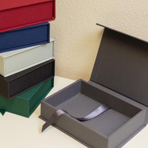 May include: A stack of colorful, rectangular gift boxes in red, blue, gray, black, and green. One box is open, revealing a gray interior and a ribbon. The boxes are on a white surface.