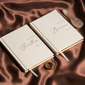 May include: Two off-white vow books with gold trim and attached ribbon bookmarks. The books are inscribed with names and dates in elegant script. A clear bottle and two gold wedding rings are also visible.