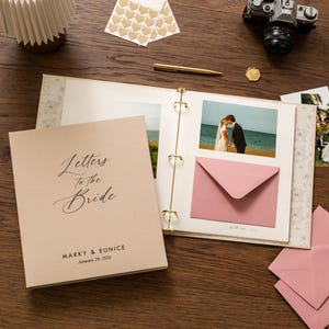 May include: A wedding-themed scrapbook with the text "Letters to the Bride" on the cover. The scrapbook is open, displaying photos, a pink envelope, and gold accents. A vintage camera and other wedding-related items are also present.
