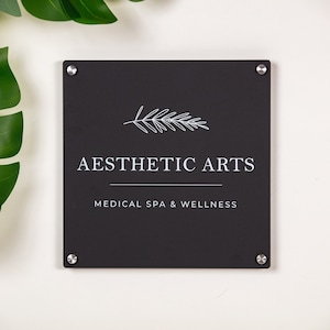 May include: A black sign with silver screws, a white stylized branch design, and the text "Aesthetic Arts Medical Spa & Wellness".
