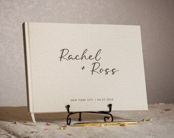 May include: A white linen guest book with the names "Rachel + Ross" and the text "NEW YORK CITY | 02.27.2022" printed in black.