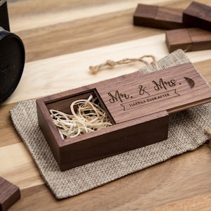 May include: A wooden box with a hinged lid, engraved with the text "Mr. & Mrs. Happily Ever After". The box is open and filled with wood shavings. The box is sitting on a burlap surface.