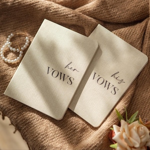 May include: Two beige vow books with the words "her vows" and "his vows" printed in black script. The books are on a textured brown surface, with a pearl bracelet and a small floral arrangement nearby. Wedding theme.