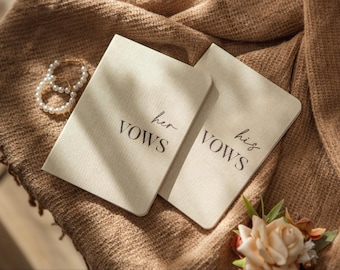 May include: Two beige vow books with the words "her vows" and "his vows" printed in black script. The books are on a textured brown surface, with a pearl bracelet and a small floral arrangement nearby. Wedding theme.