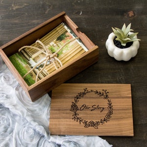May include: A rectangular wooden box with a sliding lid featuring a black engraved floral wreath and the text Our Story. Inside the box is a stack of photographs tied with jute twine. The box is made of dark brown wood with visible grain.