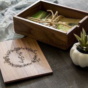 May include: A rectangular wooden box with a removable lid featuring a laser-engraved floral wreath and the text Our Story. The open box contains a stack of photographs tied with a natural jute twine cord, accompanied by a small succulent plant.