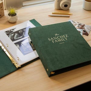 May include: A dark green photo album with gold lettering that reads "The Sanchez Family 2026" sits open next to a closed album. The open album displays photos behind clear plastic sleeves. A wooden clock, camera, and pens are in the background.