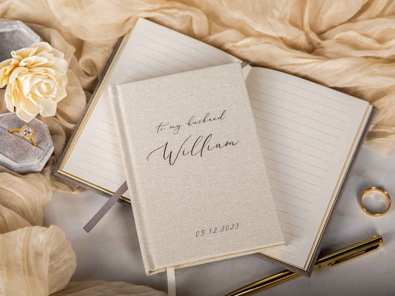 May include: A cream-colored journal with the text "to my husband William" and the date 05.12.2023. The journal is open, revealing lined pages. A gold pen and wedding ring are also visible.