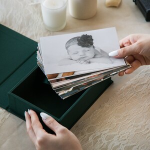 May include: A green fabric-covered box with a stack of black and white photos of a baby inside. The top photo shows a baby girl sleeping with a flower headband.