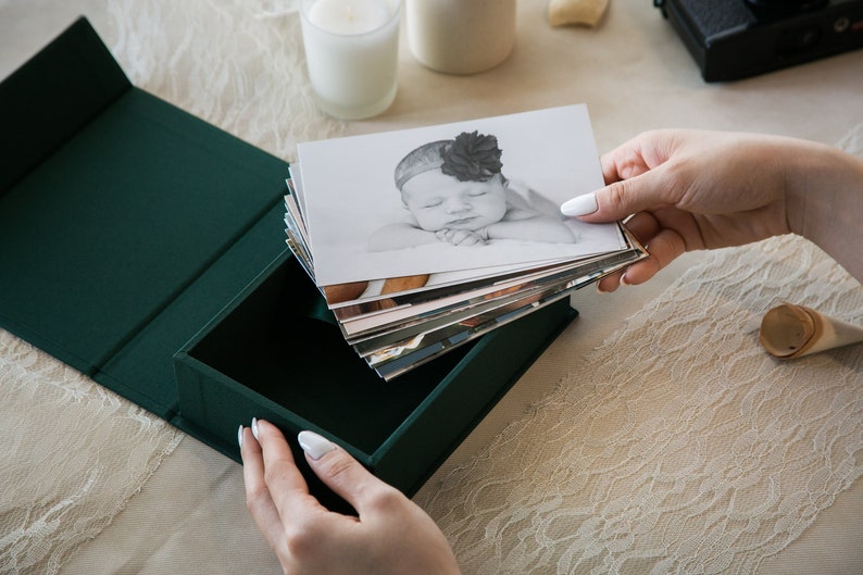 May include: A green fabric-covered box with a stack of black and white photos of a baby inside. The top photo shows a baby girl sleeping with a flower headband.
