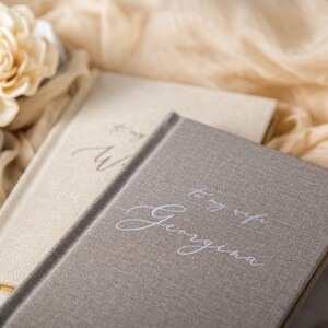 May include: Two hardcover journals with linen-like covers. One is light beige, the other is gray. The gray journal has the inscription "To my wife Georgina" in elegant script. A gold ring in a gray velvet box and a cream-colored flower are in the background.