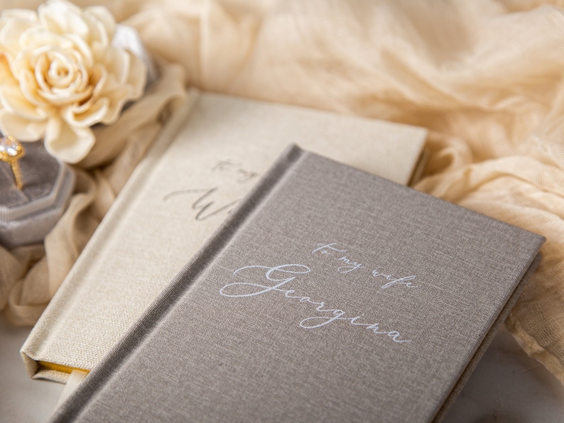 May include: Two hardcover journals with linen-like covers. One is light beige, the other is gray. The gray journal has the inscription "To my wife Georgina" in elegant script. A gold ring in a gray velvet box and a cream-colored flower are in the background.