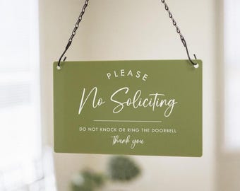 May include: An olive green rectangular sign with white script reading "PLEASE No Soliciting". Beneath, it states "DO NOT KNOCK OR RING THE DOORBELL Thank you". The sign is suspended by a black chain.