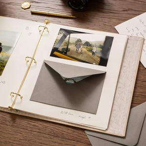 May include: An open scrapbook with photos, a gray envelope, and a handwritten note on a wooden surface. The scrapbook has gold-colored rings and a floral patterned border. A gold pen and a coin are also visible.