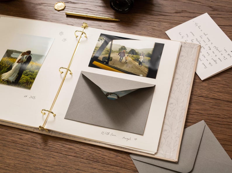 May include: An open scrapbook with photos, a gray envelope, and a handwritten note on a wooden surface. The scrapbook has gold-colored rings and a floral patterned border. A gold pen and a coin are also visible.
