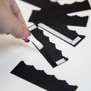 Puede incluir: Una mano sujetando un rollo de cinta adhesiva blanca sobre una tarjeta de papel negro festoneada. La tarjeta tiene una tira adhesiva blanca en la parte posterior.