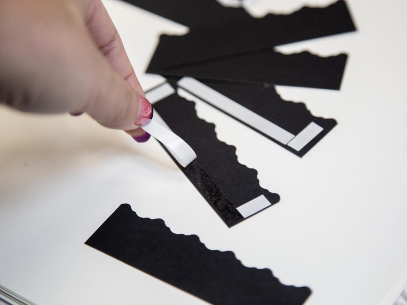 Puede incluir: Una mano sujetando un rollo de cinta adhesiva blanca sobre una tarjeta de papel negro festoneada. La tarjeta tiene una tira adhesiva blanca en la parte posterior.