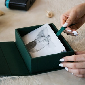 May include: A green box with a photo album inside. The album is open to a black and white photo of a baby sleeping. The box has a ribbon pull.