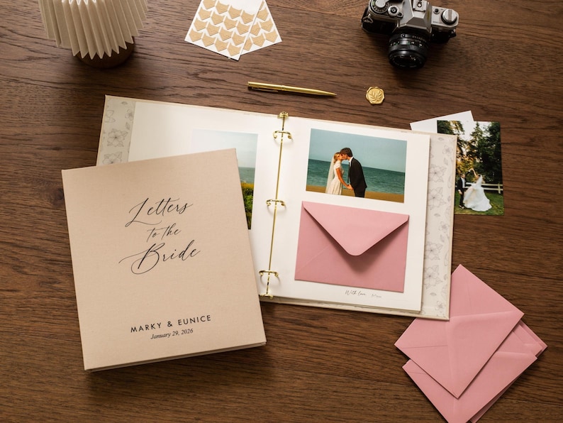 May include: A wedding keepsake album with the words "Letters to the Bride" on the cover. The album is open, displaying photos and a pink envelope. A vintage camera, pen, and stickers are also on the wooden surface.